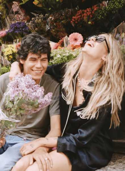 Young couple sitting at a flower market, smiling and laughing, holding lilac flowers.