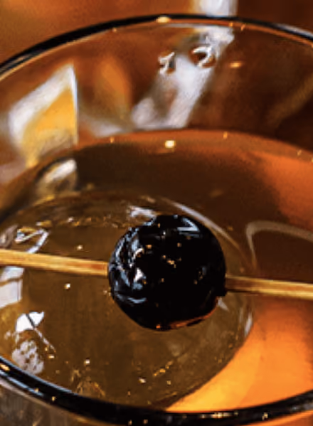 Close-up of a cocktail glass with an ice cube and a dark cherry skewered on a thin wooden stick resting on the glass rim, with warm ambient lighting reflected on the surface.