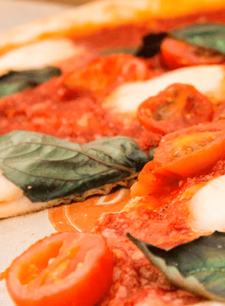 Close-up of a Margherita pizza showing slices of cherry tomatoes, fresh basil leaves, melted mozzarella cheese, and tomato sauce on a thin crust.