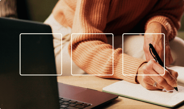 Person in a peach-colored sweater writing with a pen on a notebook, next to a partially visible laptop on a wooden surface.