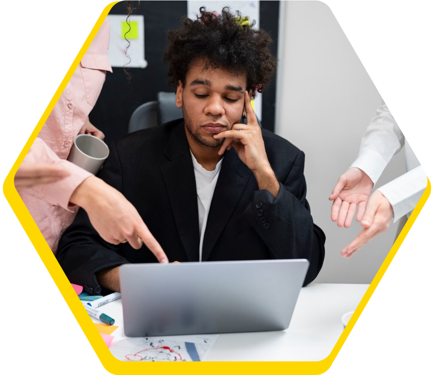 Tired man in a black blazer sitting at a laptop with people around him pointing and gesturing.