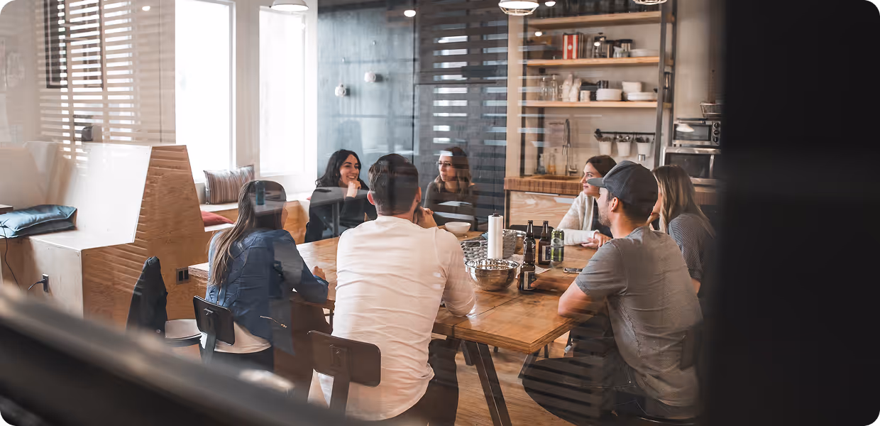 Seven people sitting around a wooden table in a modern office kitchen, engaged in a discussion.