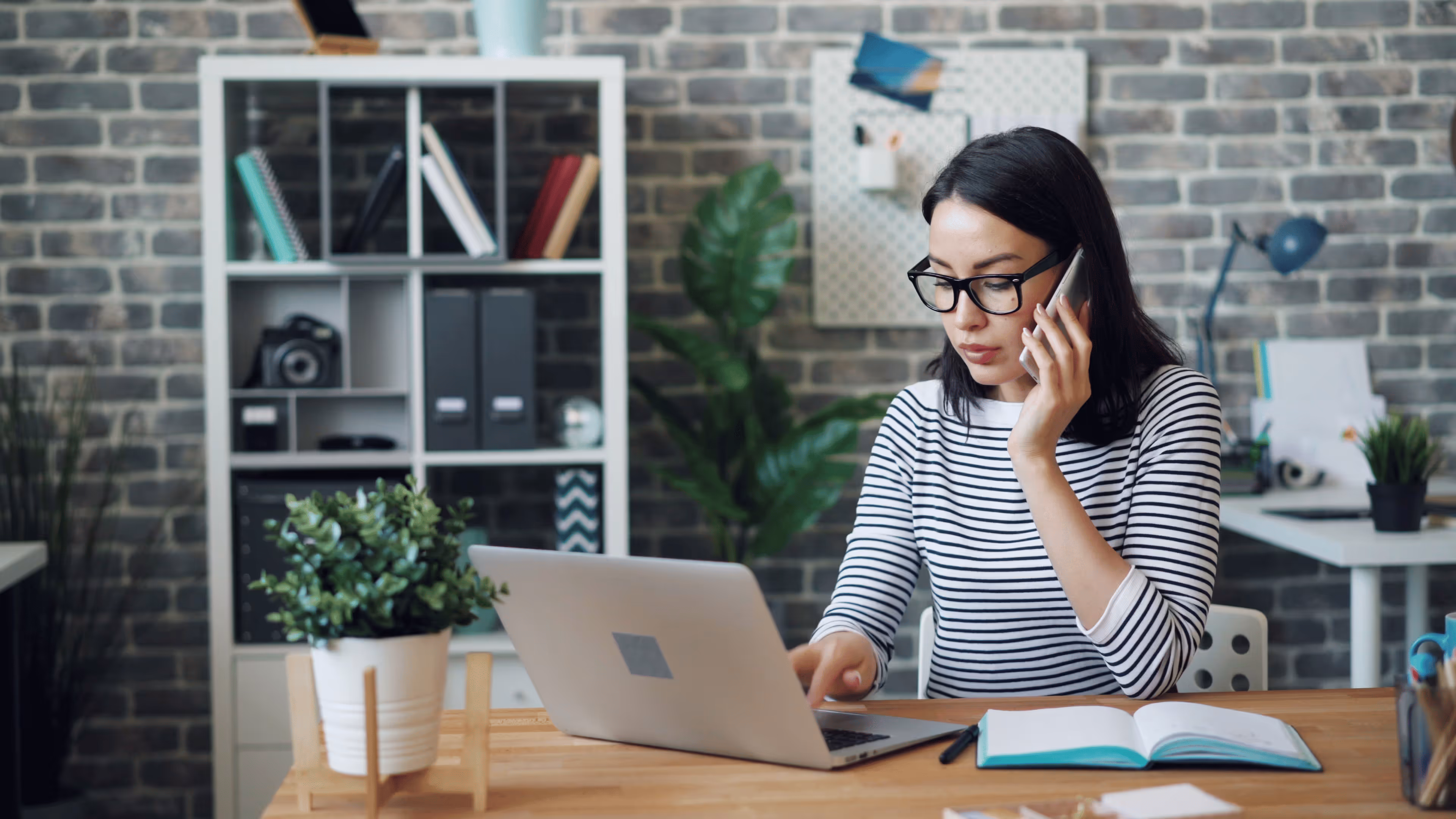 Woman with glasses talking on a smartphone while working on a laptop at a wooden desk with a notebook and plant.