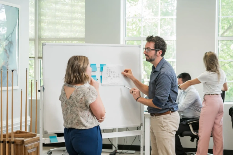 Man presenting a diagram on paper to a woman in front of a whiteboard in a bright office with two other colleagues working in the background.