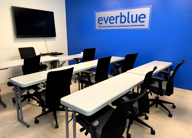 Classroom with white tables, black chairs, a wall-mounted TV, and a blue wall featuring an Everblue sign that reads 'Training Earth's Sustainable Workforce'.