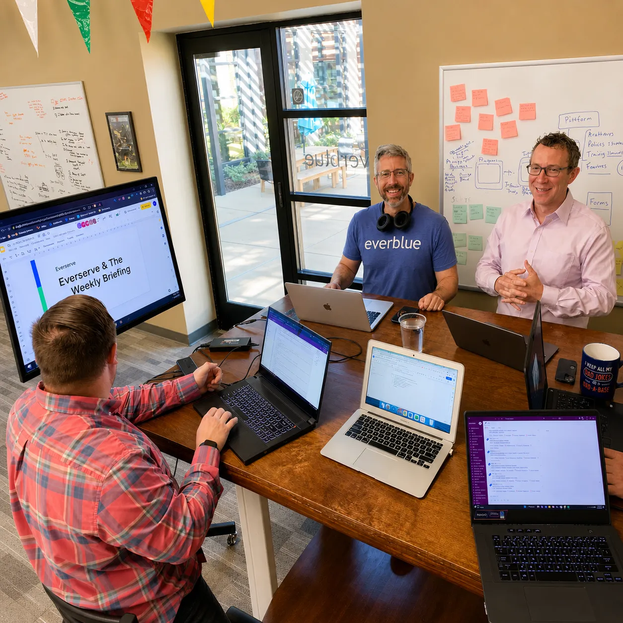 Three men having a meeting around a table with laptops and a screen displaying 'Everserve & The Weekly Briefing' in a brightly lit office.