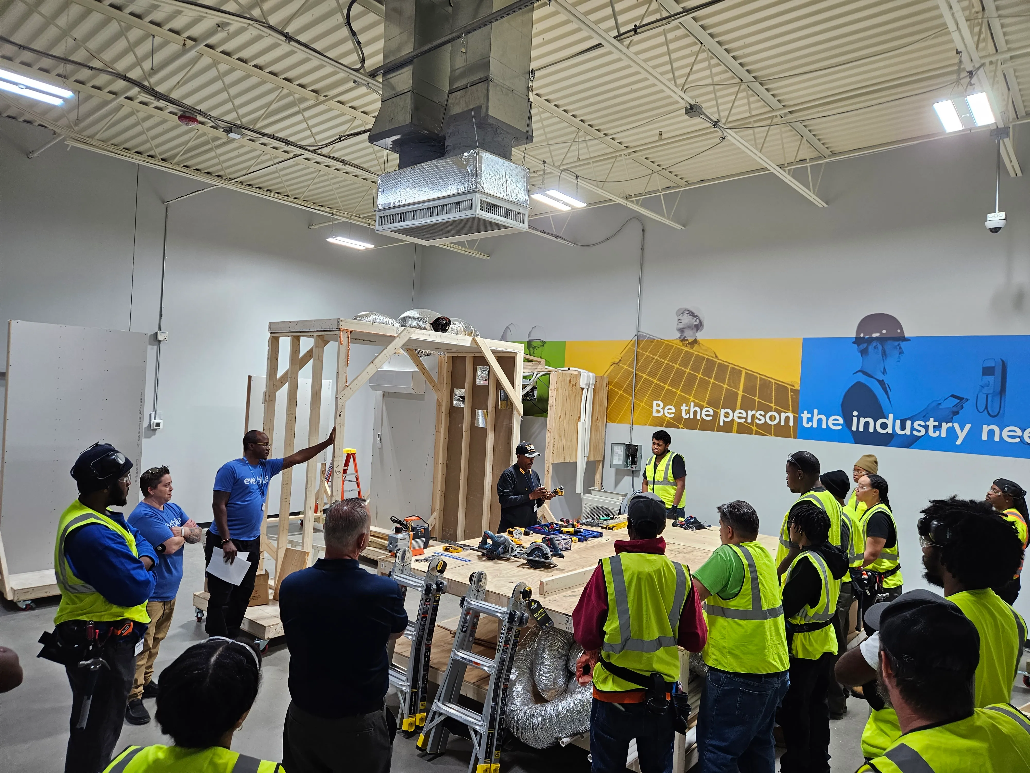 A group of people in safety vests gathered around a wooden construction frame inside a workshop, listening to an instructor.