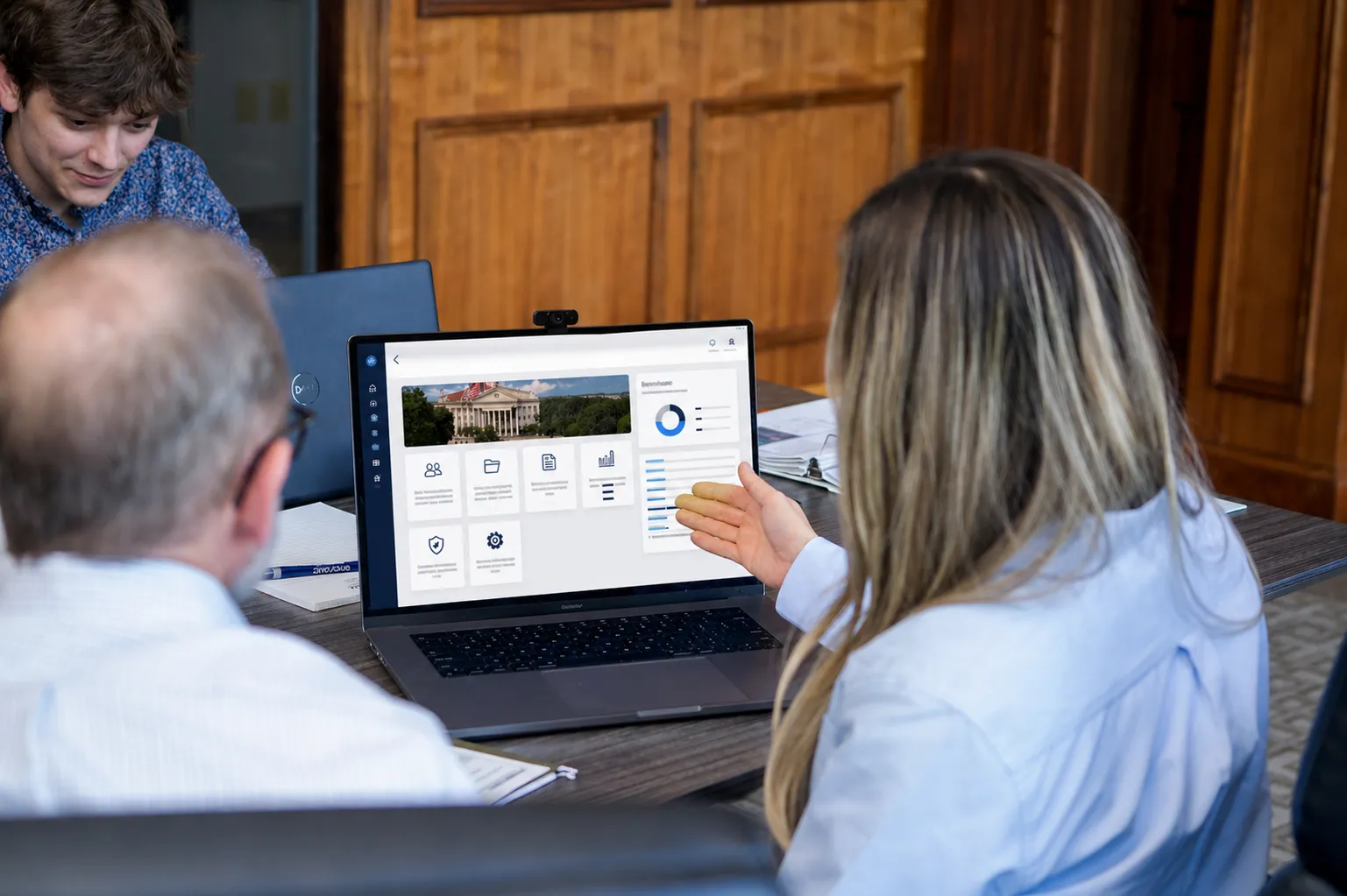 Three people in a meeting looking at a laptop screen displaying a dashboard with charts and icons.