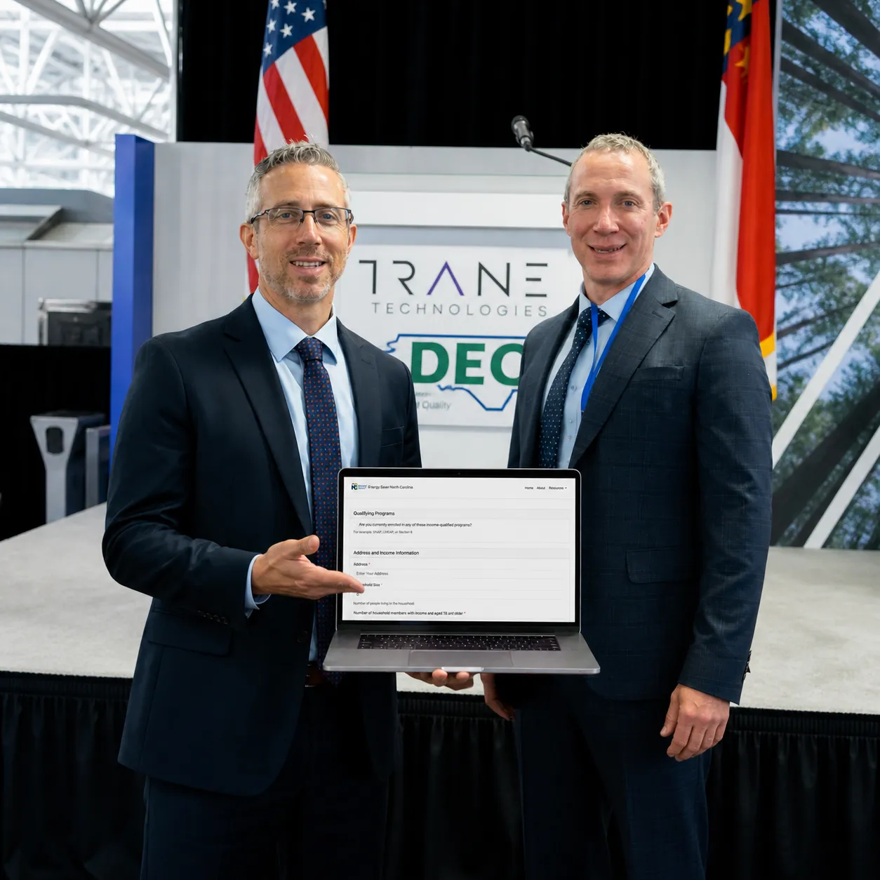 Two men in suits standing on a stage holding a laptop displaying a form, with an American flag and a North Carolina flag behind them.
