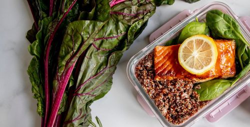 A top-down view of a personal meal container filled with quinoa, baby spinach leaves, and a slab of salmon with a lemon slice. Beside the container is a raw stock of swiss chard.