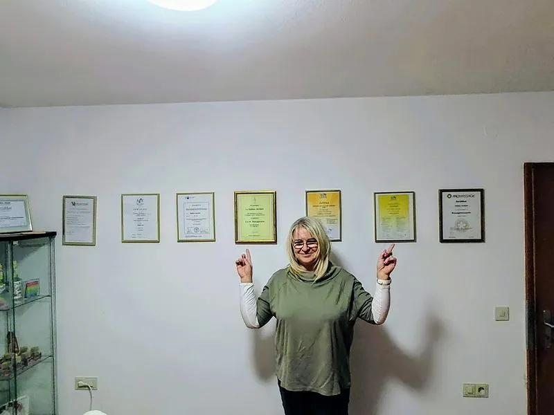 Sabine Jechow steht vor einer Wand mit gerahmten Zertifikaten und weist stolz auf ihre fachlichen Qualifikationen hin – ein klares Zeichen für ihre Expertise im Bereich Heilpraktiker Coaching, Mindset Coaching und Lebensberatung. Diese authentische Darstellung unterstreicht ihre Kompetenz für ganzheitliches Coaching und Beratung in den Heilberufen.