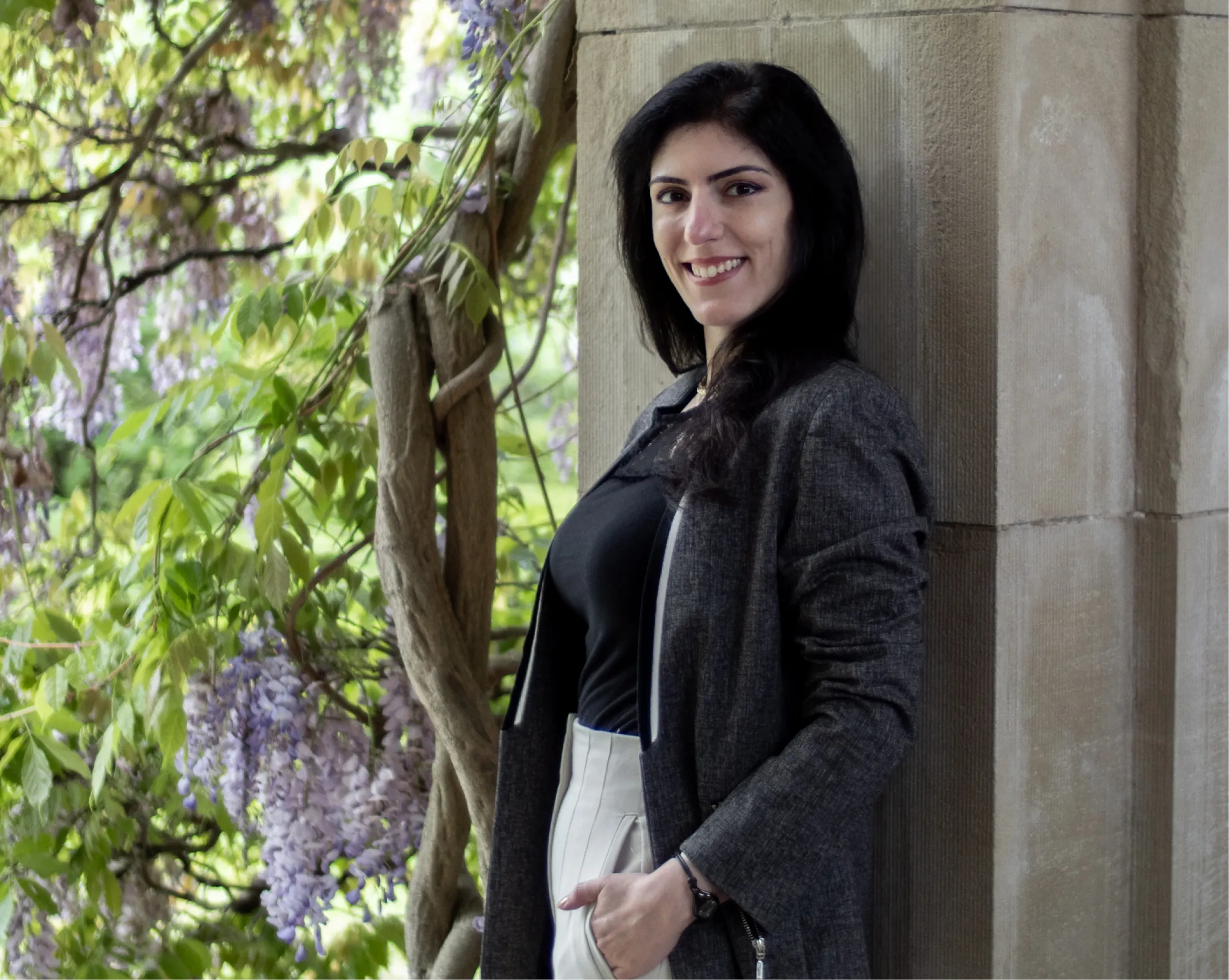 Dr. Irina Gulerez smiling against a stone wall, surrounded by greenery and purple flowers.