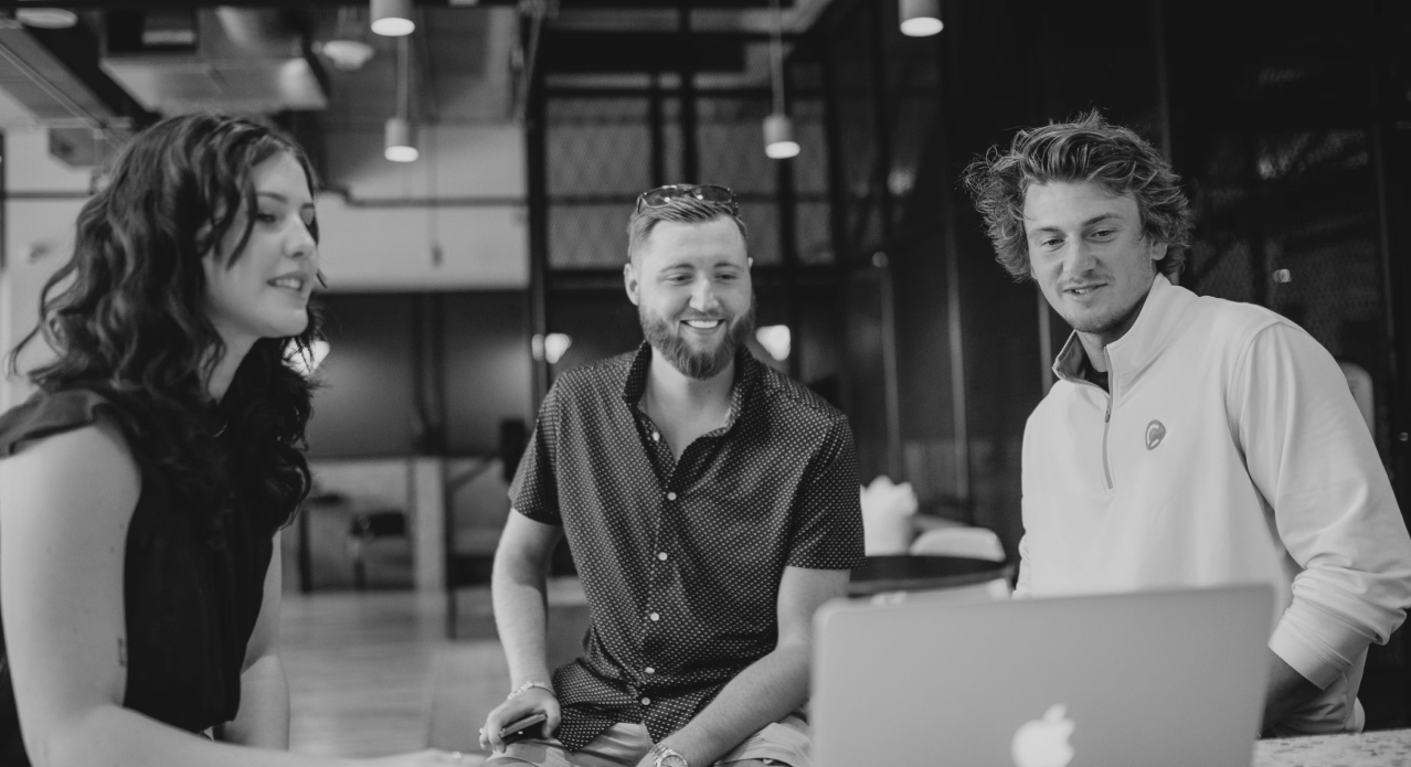 Three team members collaborating around a laptop in a modern office environment.