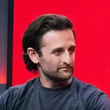Man with dark hair and beard wearing a dark gray t-shirt sitting against a red and black background.
