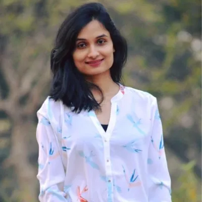 Smiling woman with long dark hair wearing a white blouse with a bird pattern standing outdoors with blurred trees in the background.