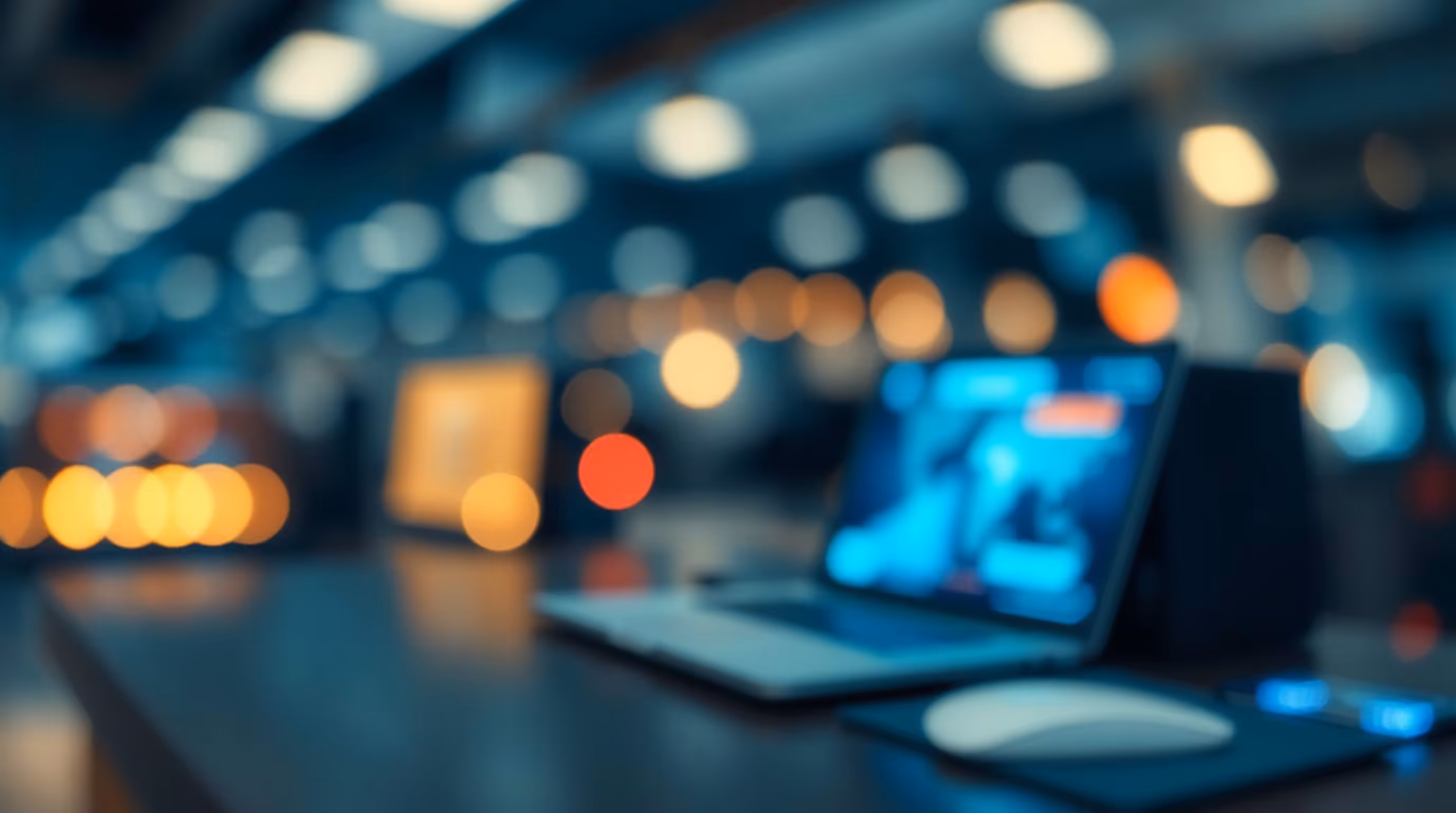 Blurred image of a laptop and mouse on a desk with warm bokeh lights in the background.