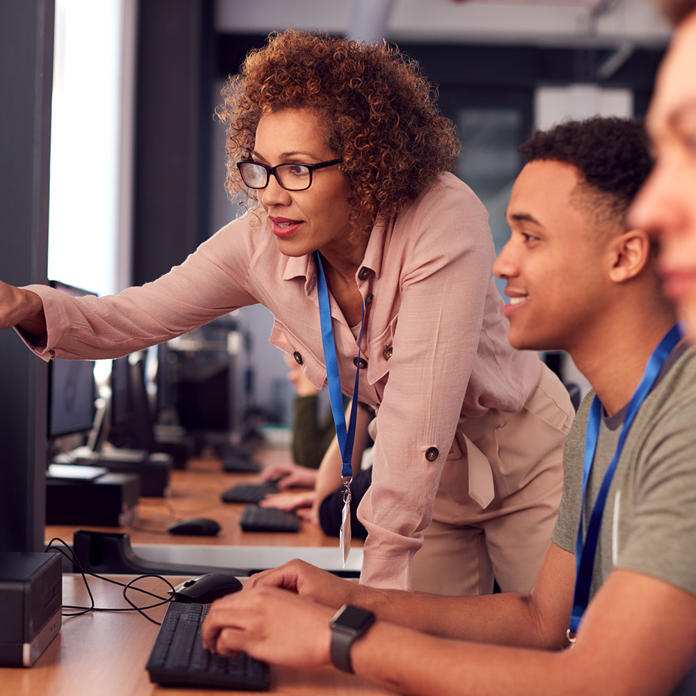 Woman with curly hair and glasses guiding a young man at a computer in a classroom setting.