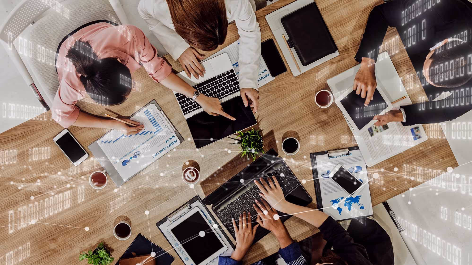 Overhead view of four people collaborating at a table with laptops, tablets, charts, and coffee cups, overlaid with digital binary code graphics.