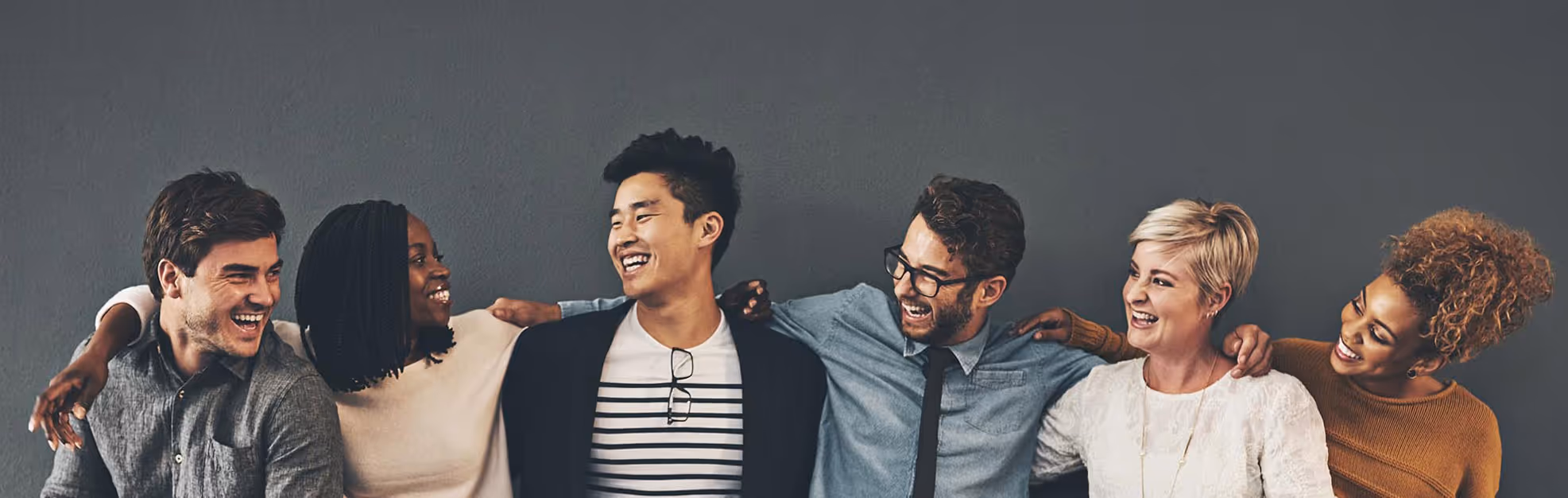 A diverse group of six friends smiling and linking arms against a gray wall.