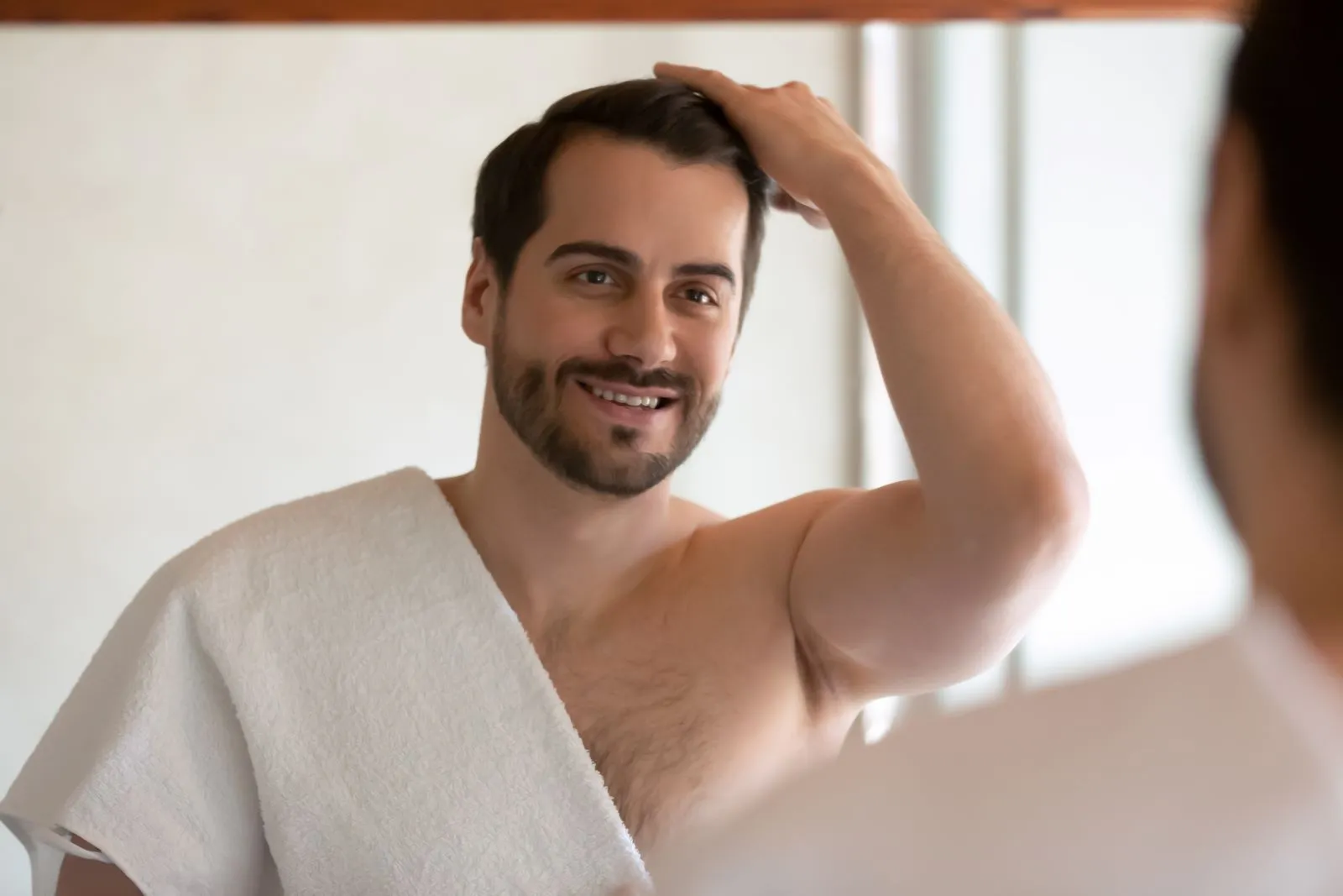 A man with a towel on his head is looking at himself in the mirror.