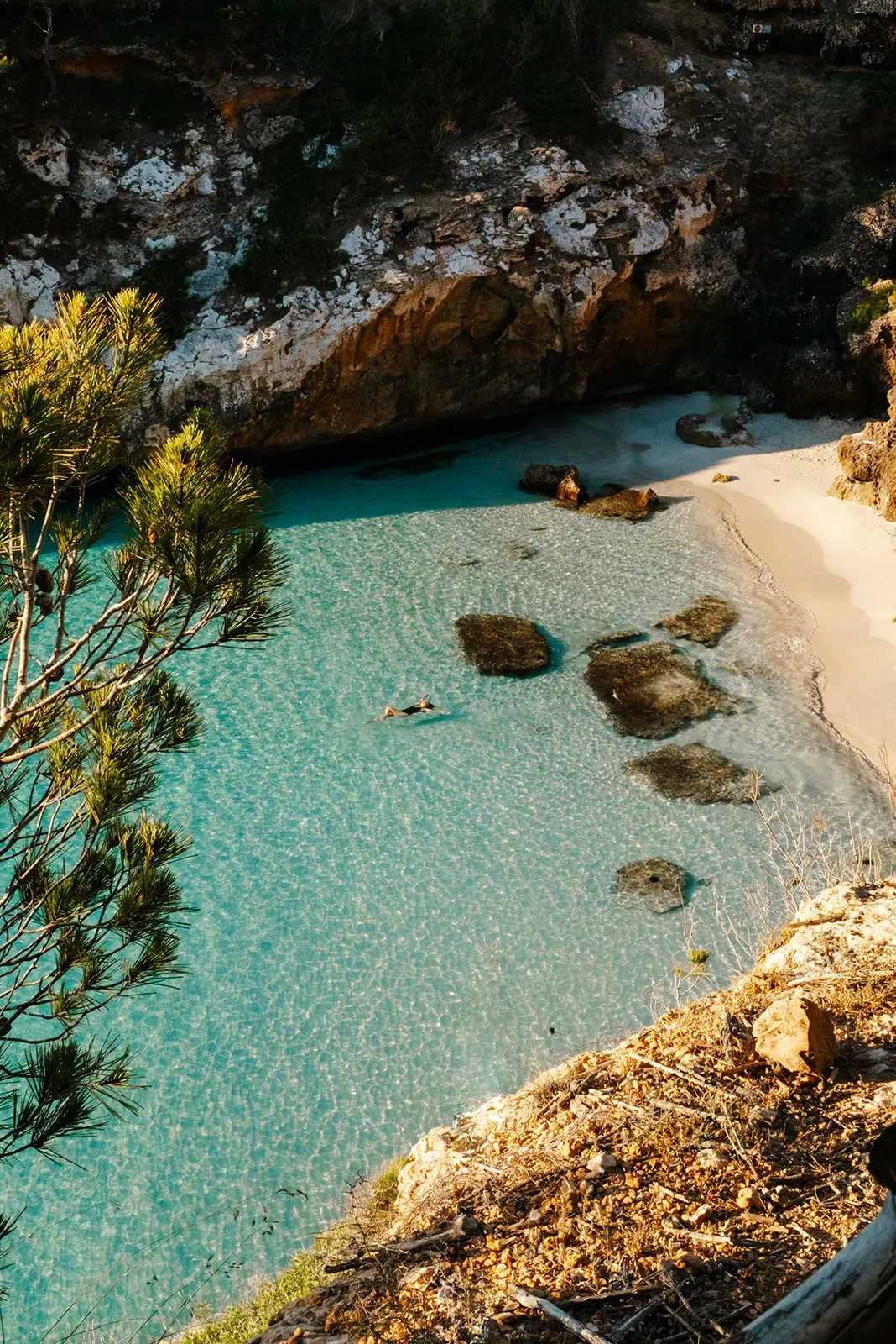 woman floating in clear turquoise water near a sandy beach cove surrounded by rocky cliffs and trees.