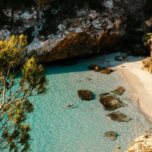 Clear turquoise water cove in Mallorca with a sandy beach, rocky cliffs, and a woman floating in the water near rocks.