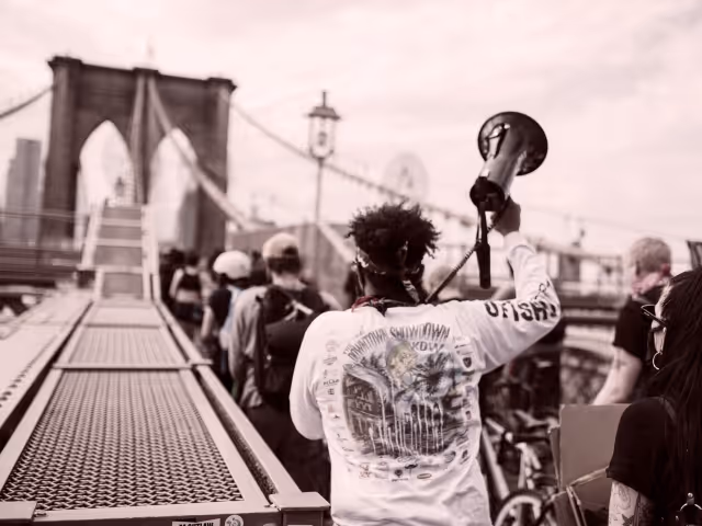 A black man speaking into a megaphone, with a group marching over the Brooklyn Bridge