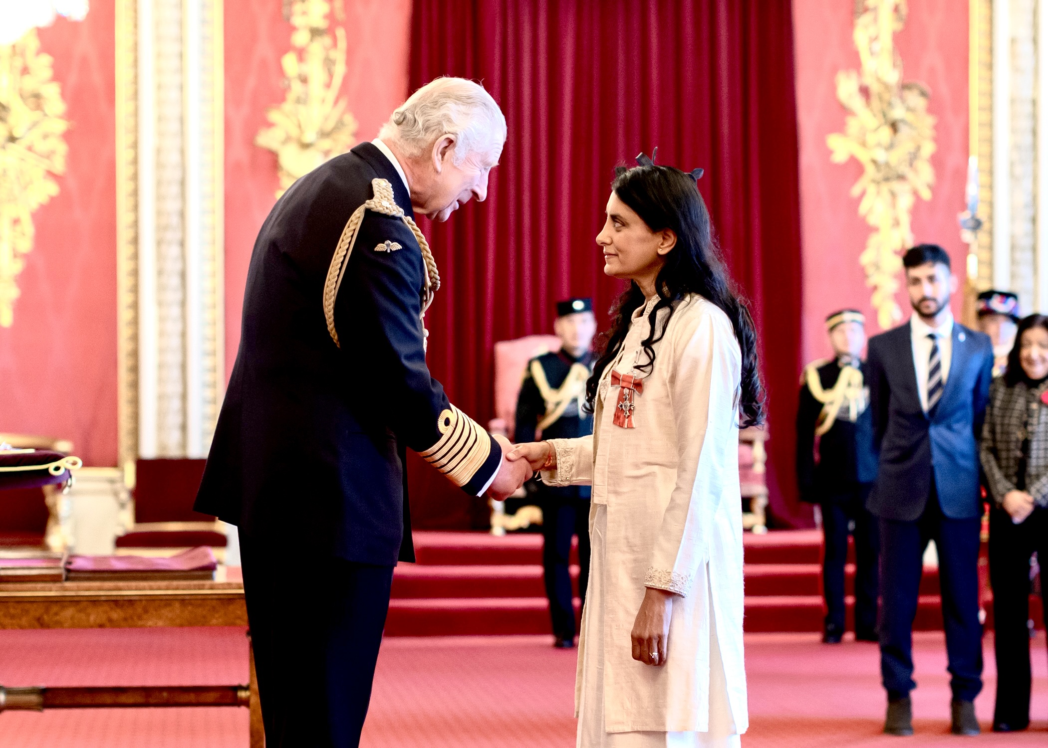 Aneeta Prem MBE at her investiture, shaking hands during the ceremony, with her mother Savita Prem and Rishi Prem Thornhill visible in the background.