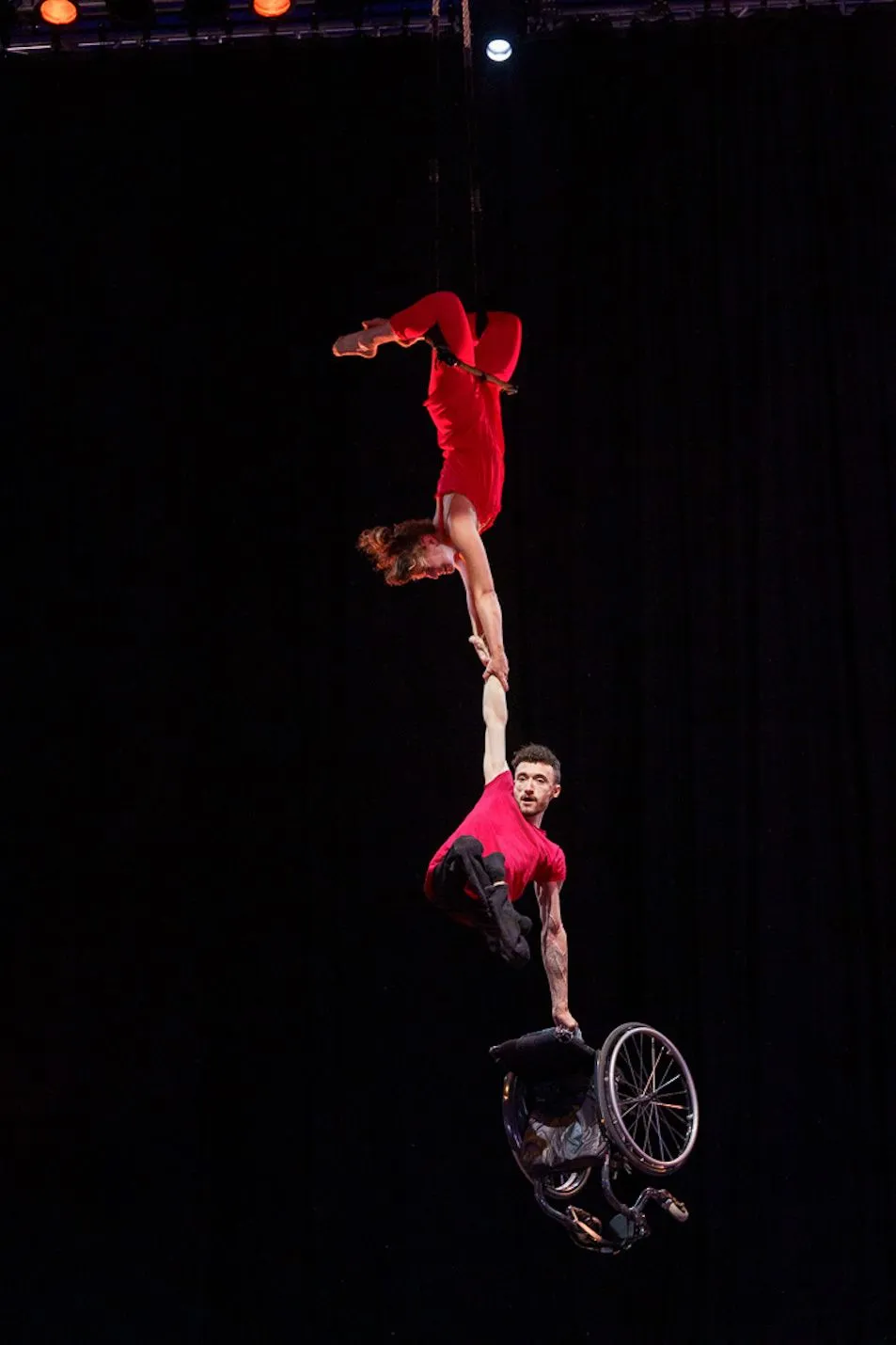 Jonny and Tilly are performing in red costumes. Jonny is hanging from Tilly's hand in the air, holding a wheelchair.