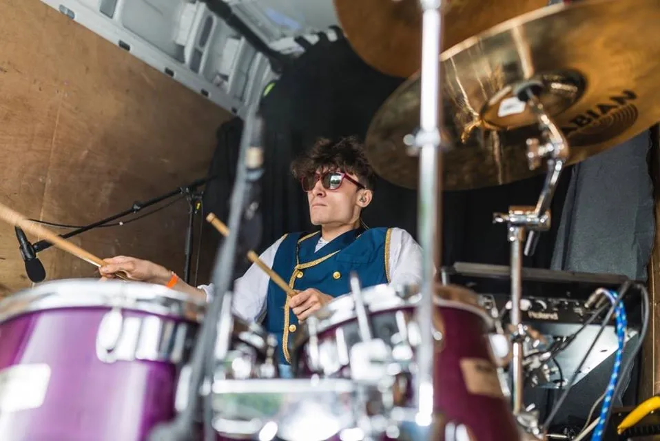 Jonny is playing drums, wearing sunglasses and holding two drumsticks.