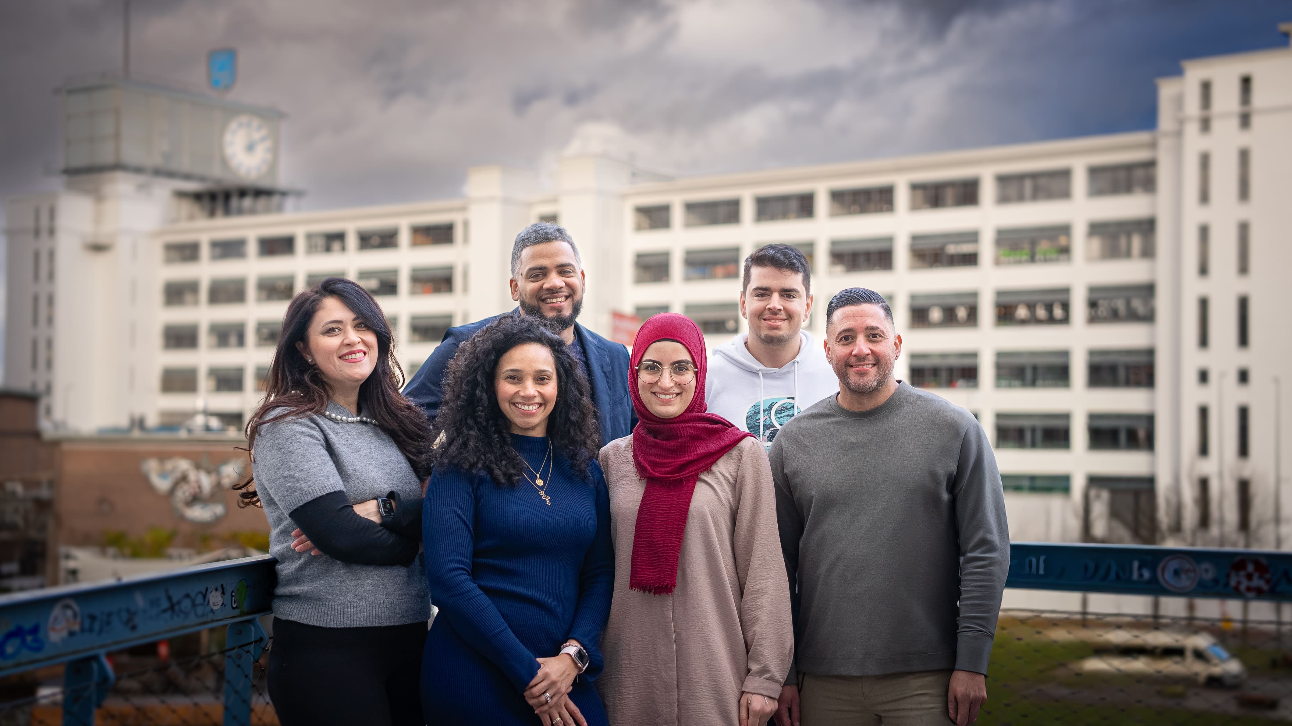 Group of six diverse young adults smiling outdoors in front of a large white building with a clock tower.
