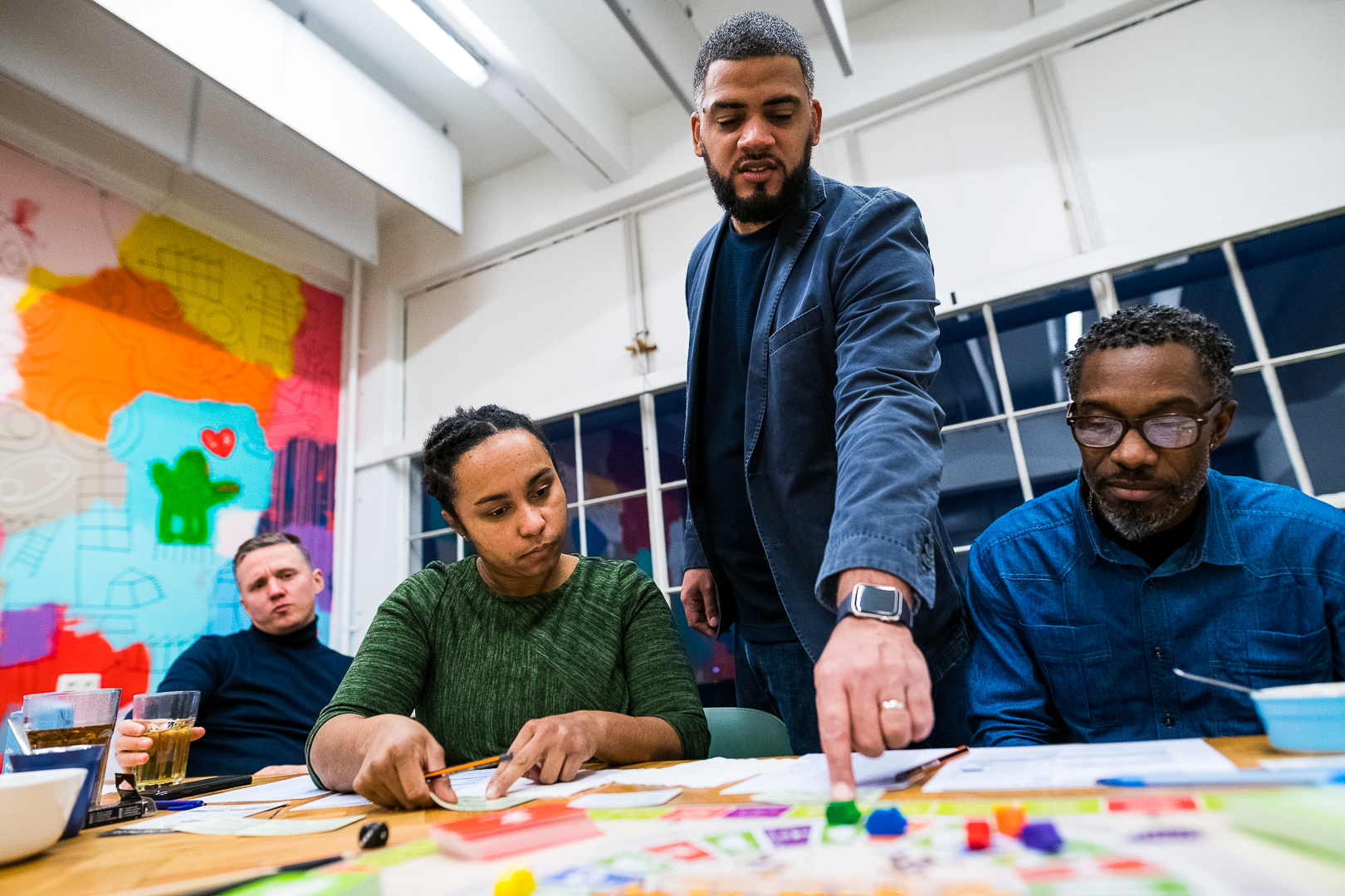 Four diverse adults playing a board game at a table in a colorful room, focused on the game pieces and cards.