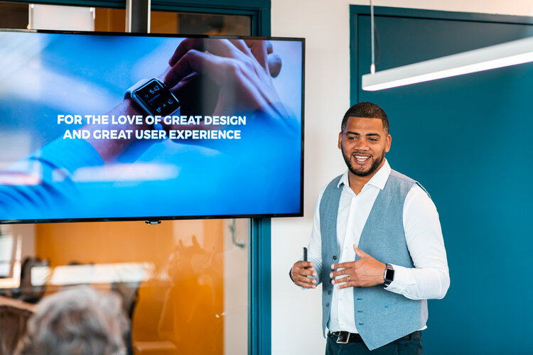 Smiling man in white shirt and gray vest giving a presentation next to a screen displaying the text 'For the love of great design and great user experience.'
