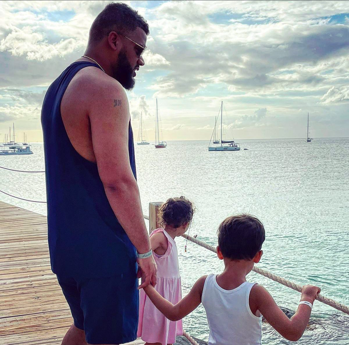 Man holding hands with two children on a wooden dock overlooking calm sea with sailboats under a partly cloudy sky.
