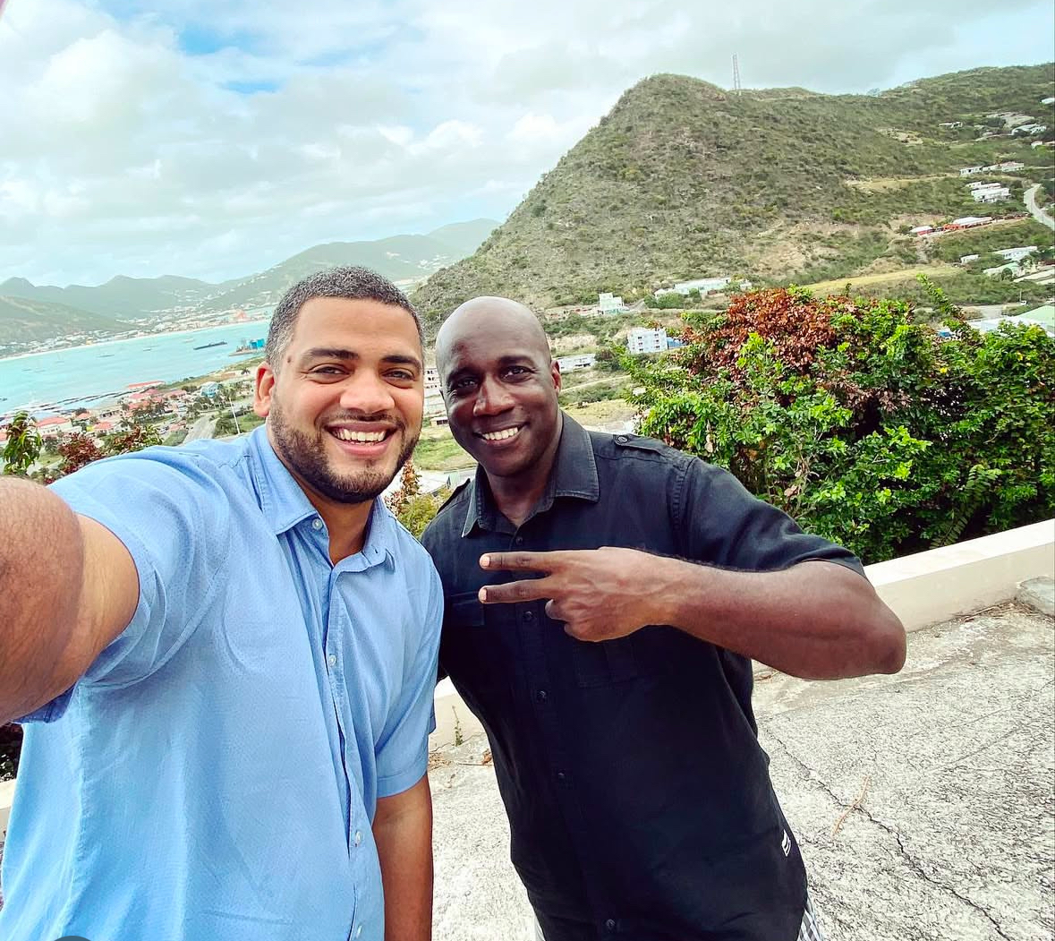 Two men smiling for a selfie outdoors with green hills and a coastal town in the background.