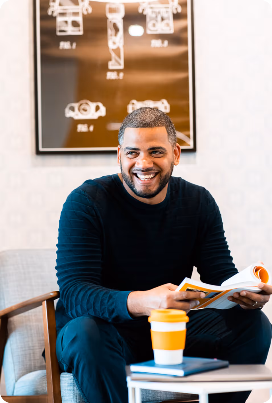 Smiling man in a black sweater sitting on a chair reading a book with a coffee cup on a table in front.