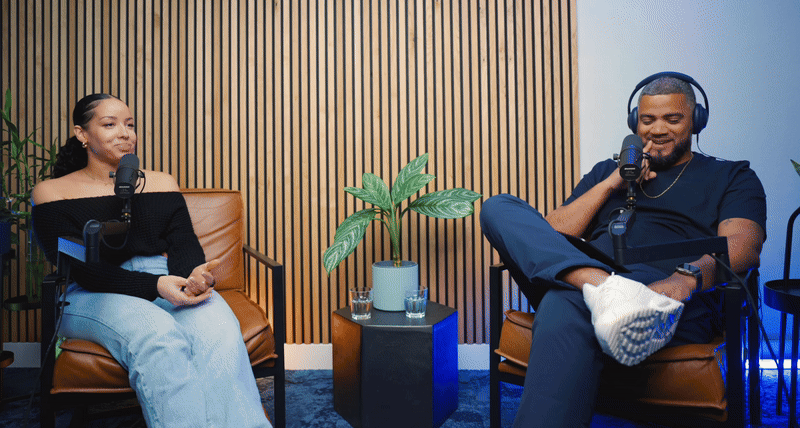 Two people sitting in brown chairs with microphones, recording a podcast in a room with a wooden slat wall and a small table with a plant between them.