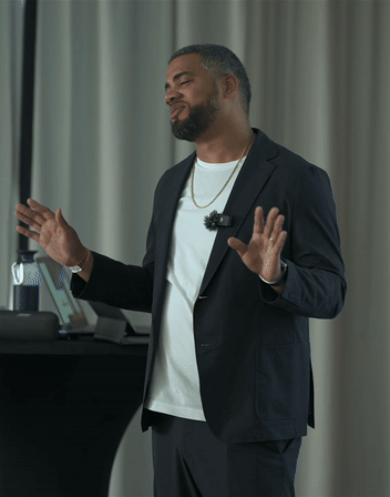 Man in black suit jacket and white shirt speaking with hands raised, standing in front of a table with a water bottle and laptop.