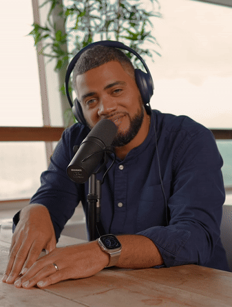 Man with headphones speaking into a microphone at a wooden table, wearing a navy blue shirt and a smartwatch.
