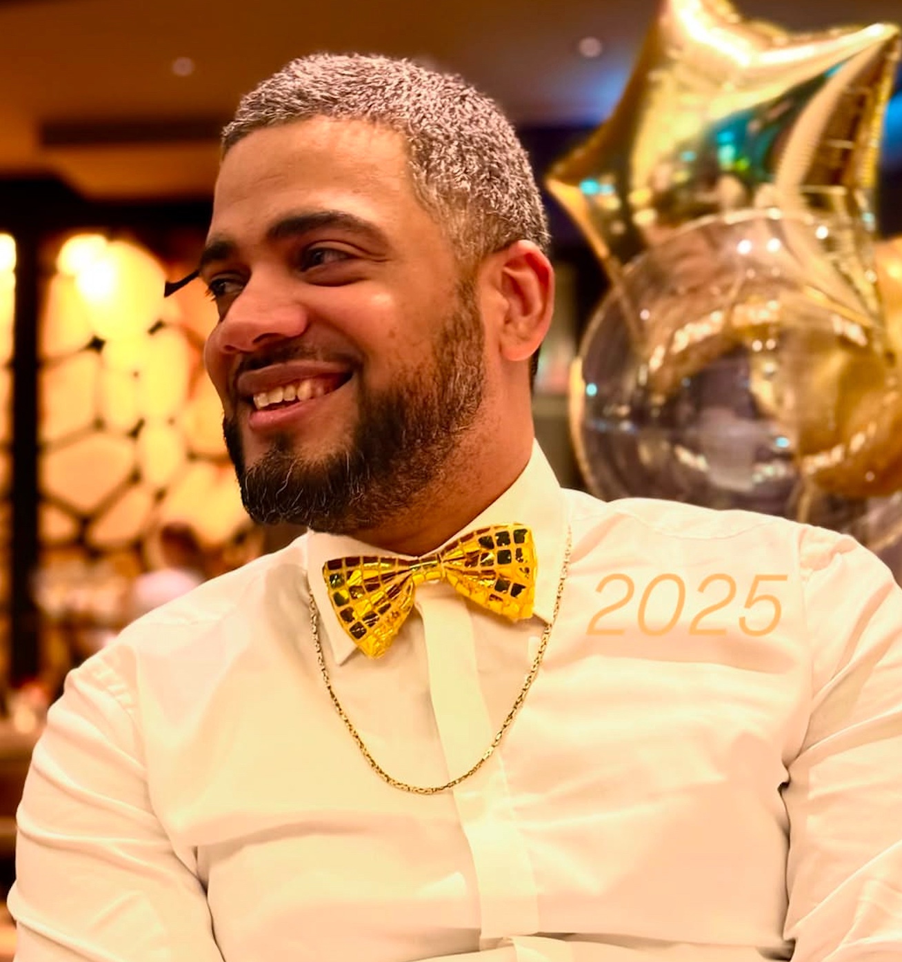 Smiling man with gray hair and beard wearing a white shirt and shiny gold bow tie, with gold star-shaped balloons in the background.