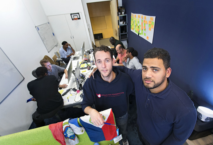 Two men posing for the camera in a busy office with six people working at computers in the background.
