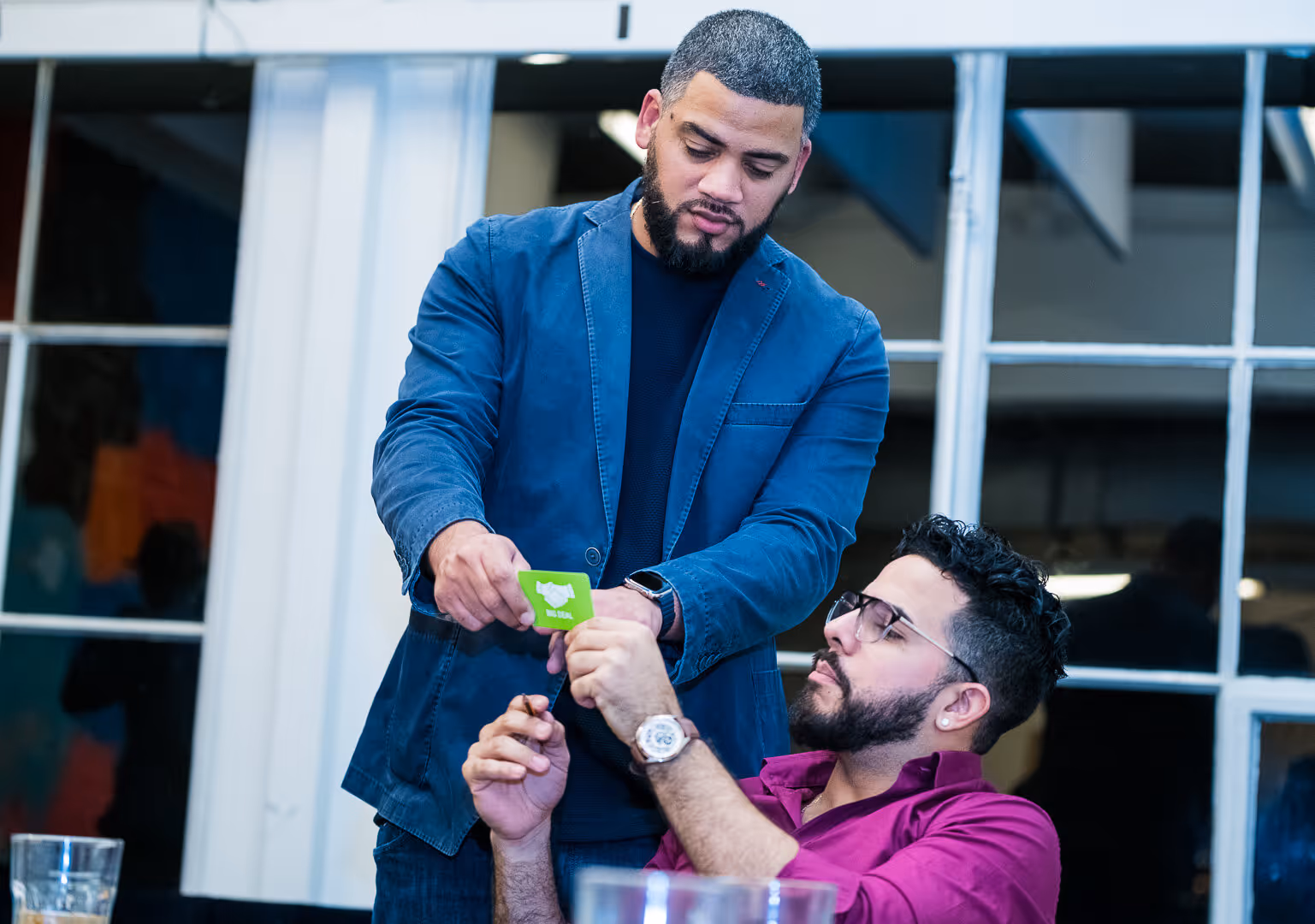 Man in blue jacket handing a green card to a seated man in a purple shirt with glasses.