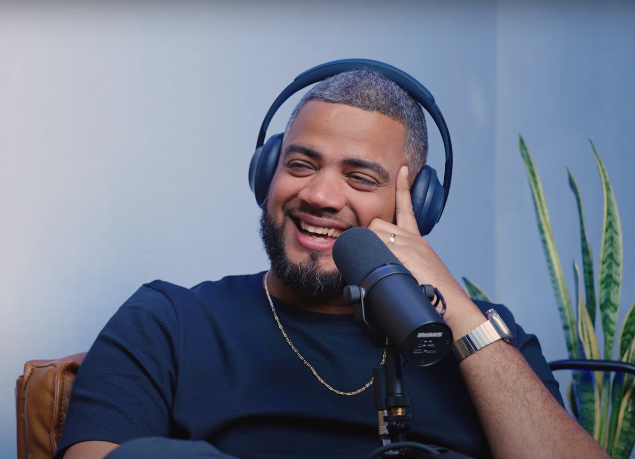 Smiling man wearing headphones speaking into a microphone with a plant in the background.