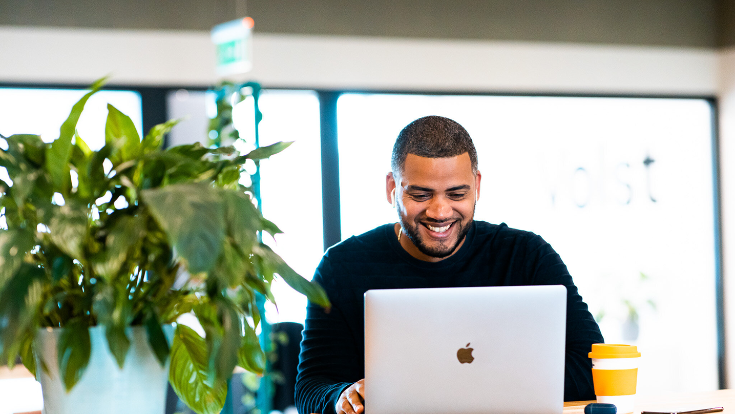 Smiling man working on a laptop at a desk with a plant and a coffee cup nearby.