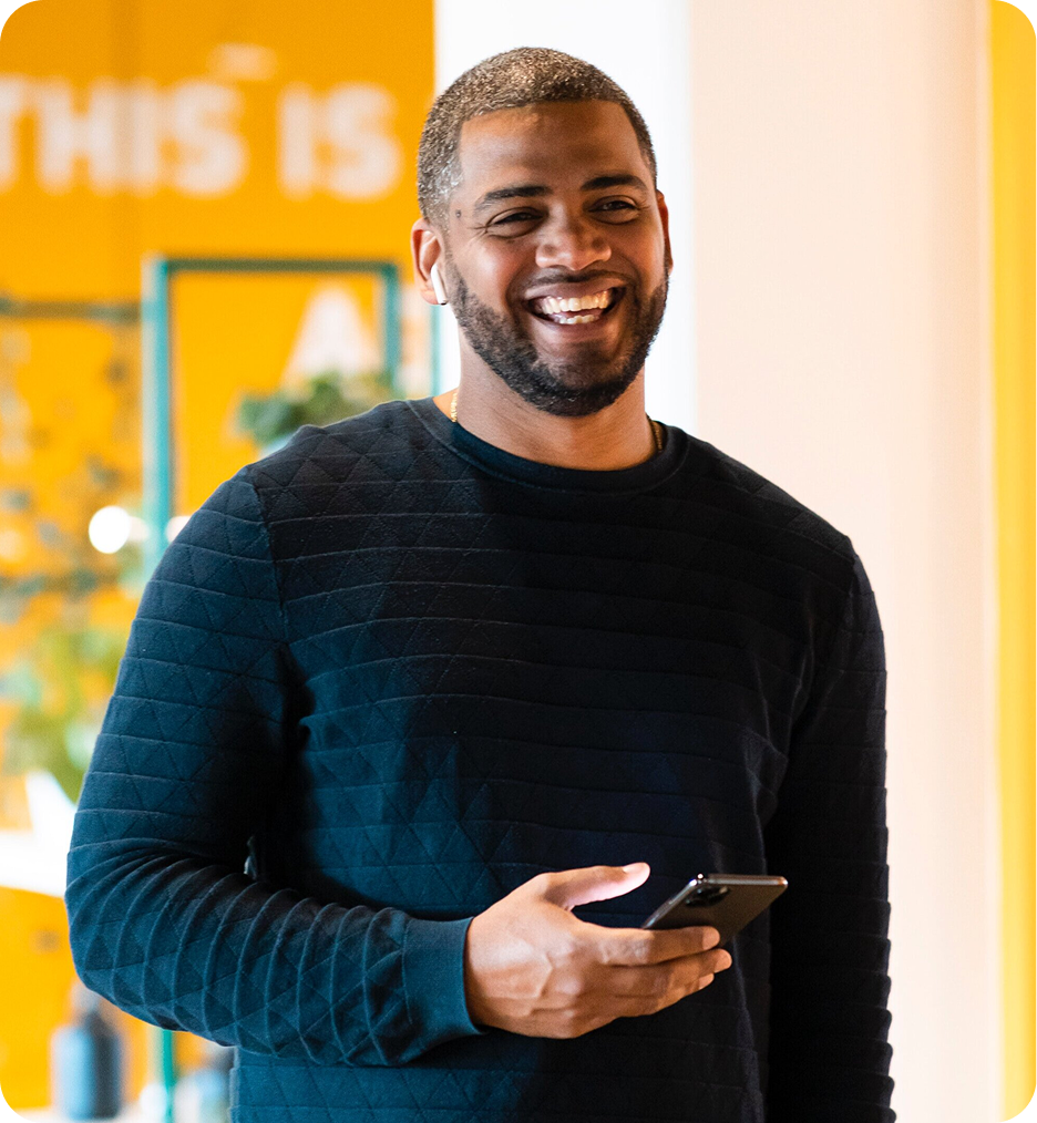 Smiling man wearing a black quilted sweater holding a smartphone indoors with a yellow background.