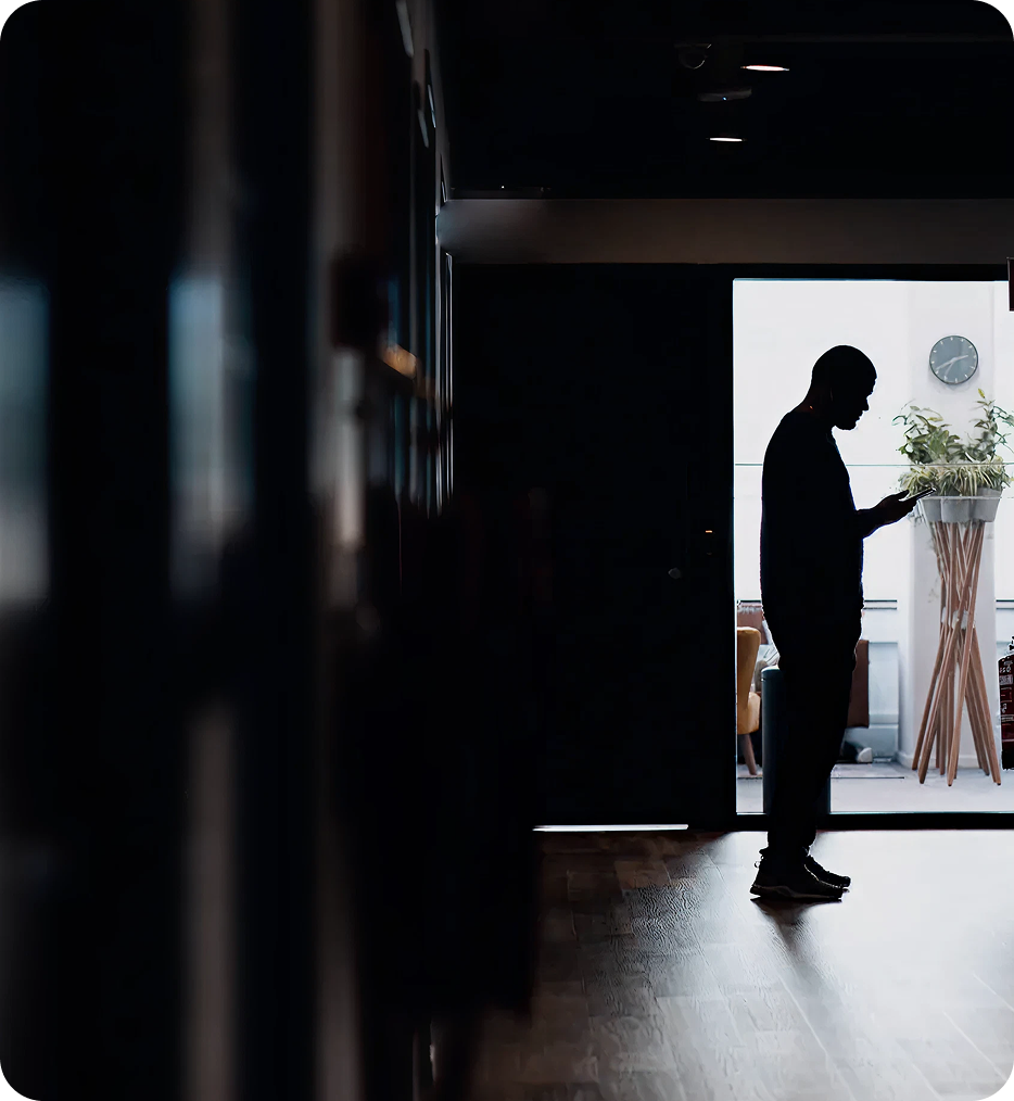 Silhouette of a person standing indoors looking at a smartphone near a bright window with a wall clock and plants in the background.