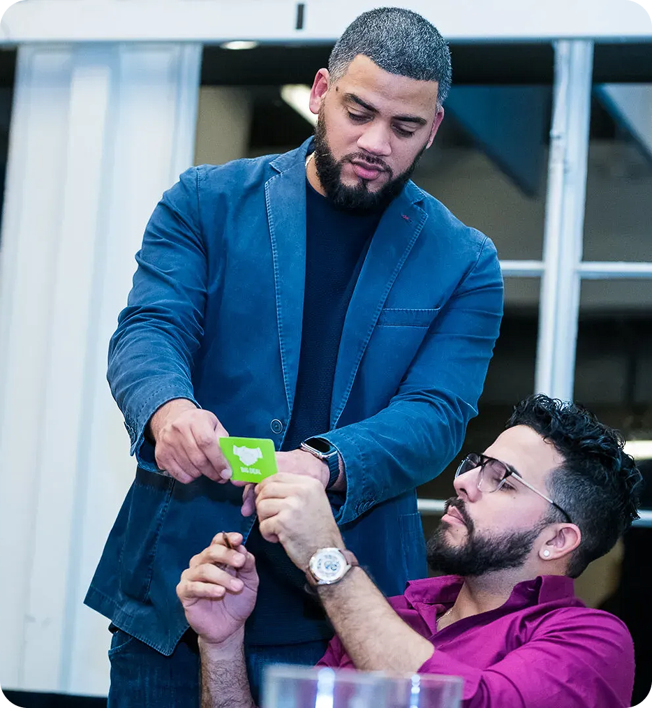 Man in blue jacket handing a green card to a seated man in a purple shirt wearing glasses.