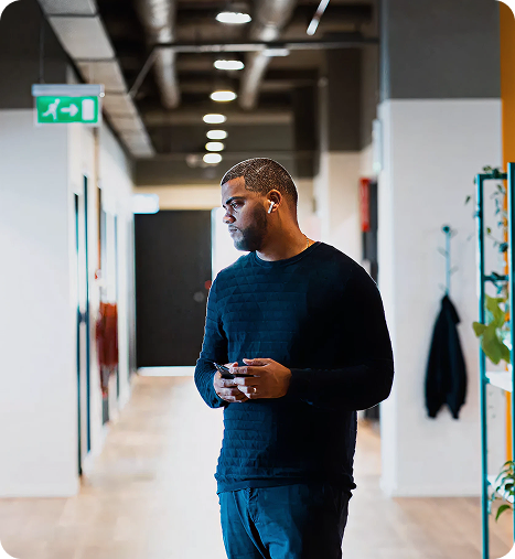 Man in a black sweater listening to music with wireless earphones and holding a phone in a modern corridor.