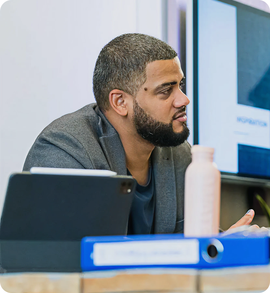 Man with short hair and beard wearing a grey blazer attentively listening in a meeting room with a tablet and blue binder on the table.