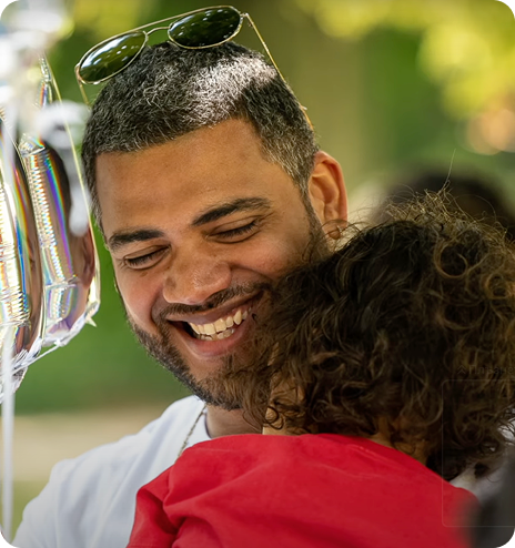 Smiling man with sunglasses on his head holding a child in a red shirt outdoors.